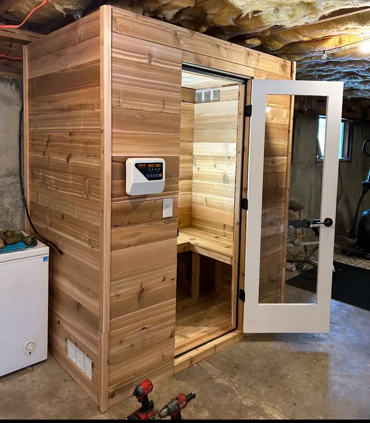 A wooden sauna with a glass door and control panel in a basement.